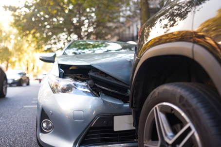 Damaged black car with a red roof off the road in a dirt ditch, showing a crushed front bumper and broken parts, surrounded by greenery.